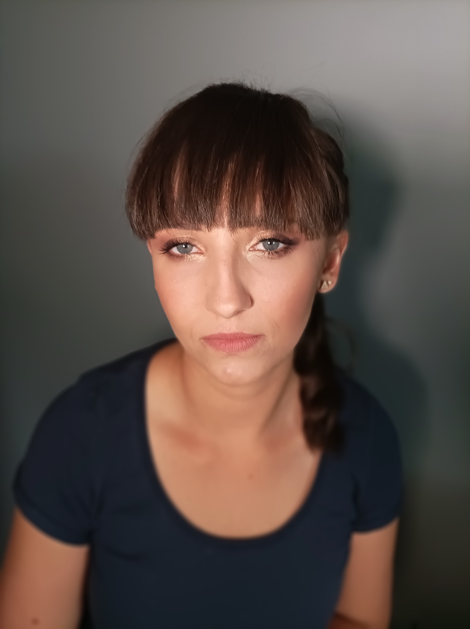 Portret kobiety z subtelnym, rozświetlającym upiększeniem oczu i delikatnym konturowaniem twarzy, włosy spięte w niski kucyk, widoczne kolczyki w kształcie trójkątów.