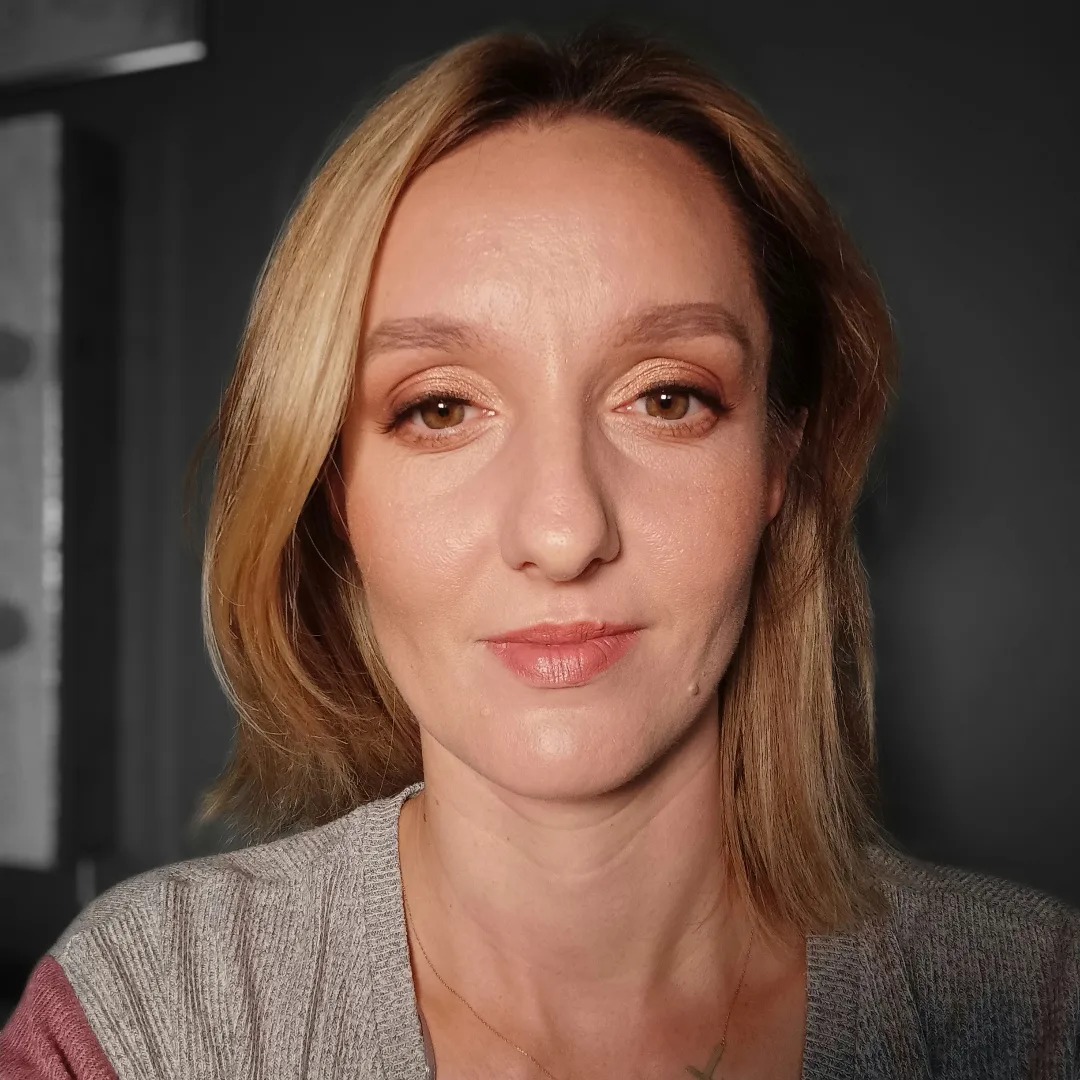 Portret kobiety z delikatnym, złotym cieniem na powiekach i subtelnie podkreślonymi ustami, włosy blond do ramion, szara bluzka.