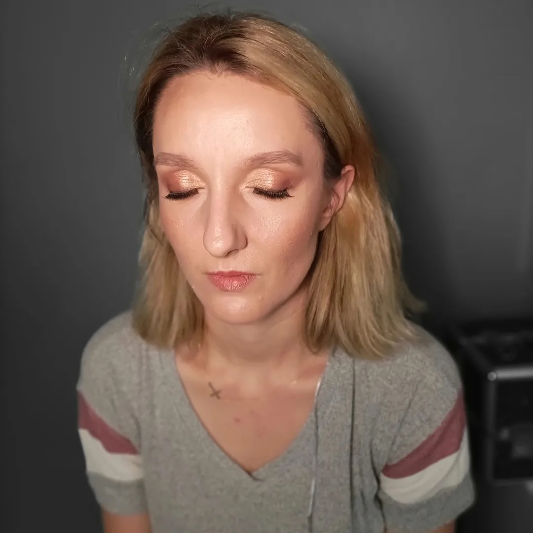 Portret kobiety z zamkniętymi oczami, prezentującej delikatny, złoty odcień cieni na powiekach, subtelny róż na policzkach i naturalny kolor ust; widoczny mały tatuaż na dekolcie.