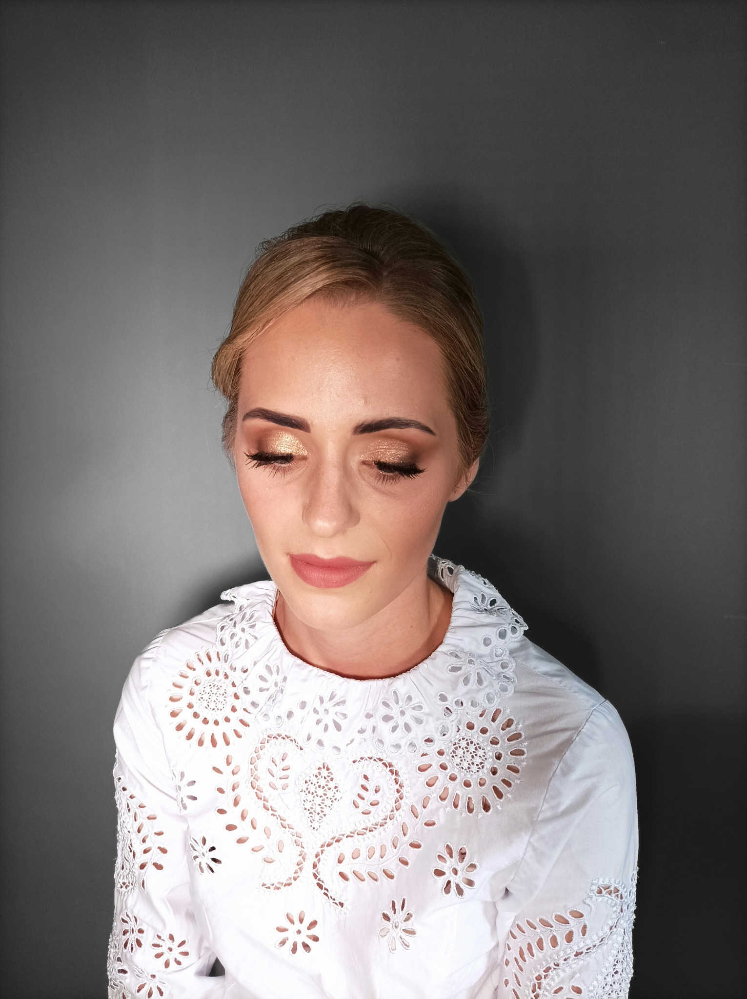 Portret kobiety z delikatnym, złotym cieniowaniem powiek i brzoskwiniowymi ustami, ubrana w białą bluzkę z ażurowymi zdobieniami.