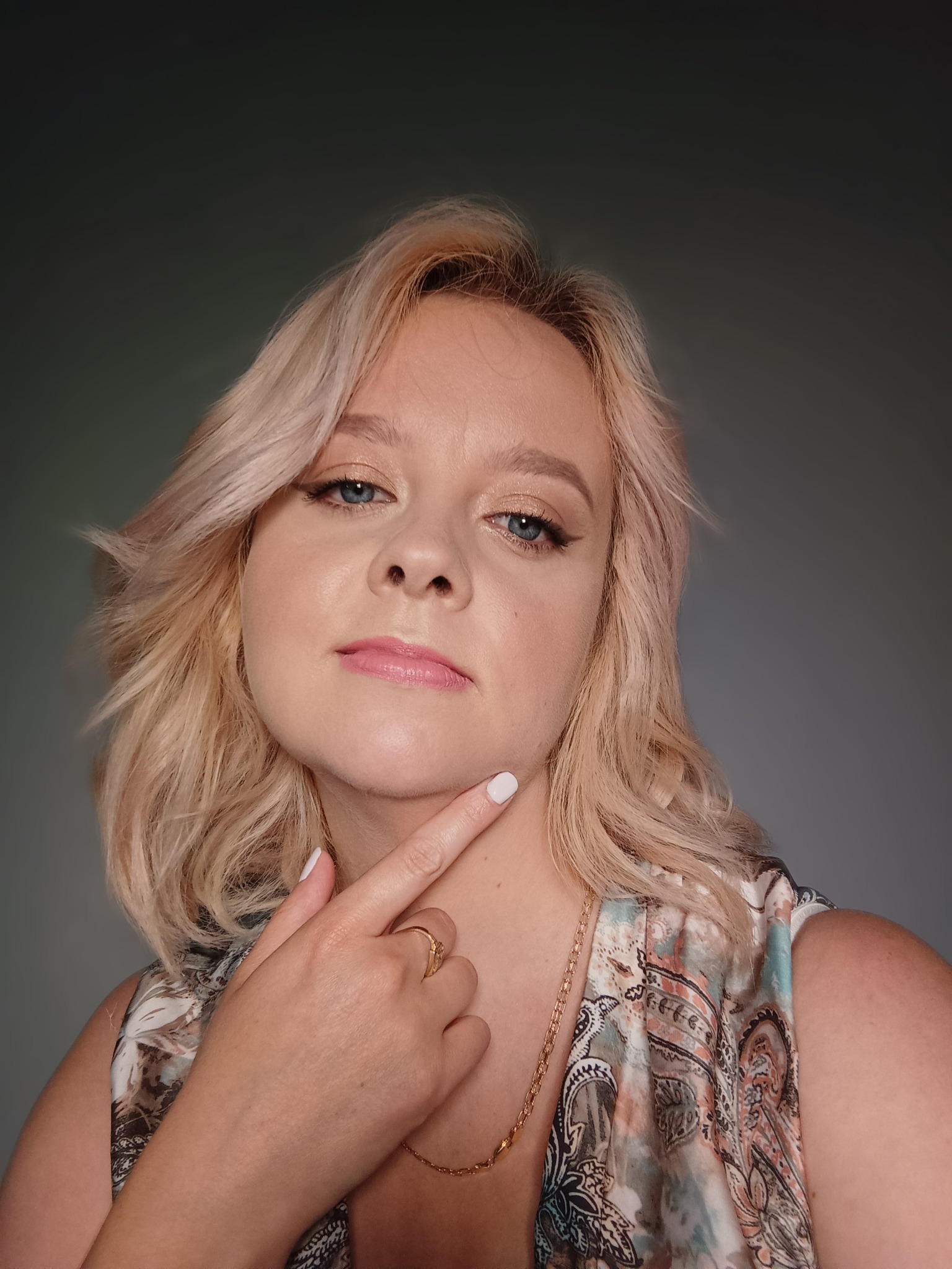 Portret kobiety z blond włosami i delikatnym podkreśleniem oka eyelinerem, wskazującej palcem na podbródek, z widocznym złotym łańcuszkiem i pierścionkiem.