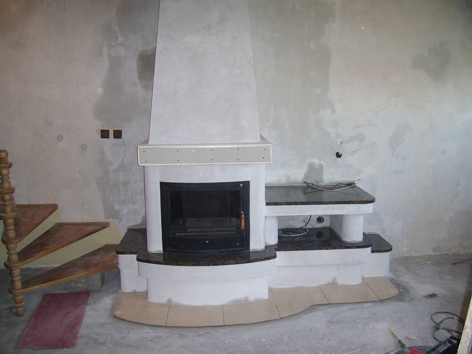 Niedokończony montaż kominka z obudową z białego betonu i blatem z ciemnego kamienia, widoczne schody drewniane i surowa ściana w tle.