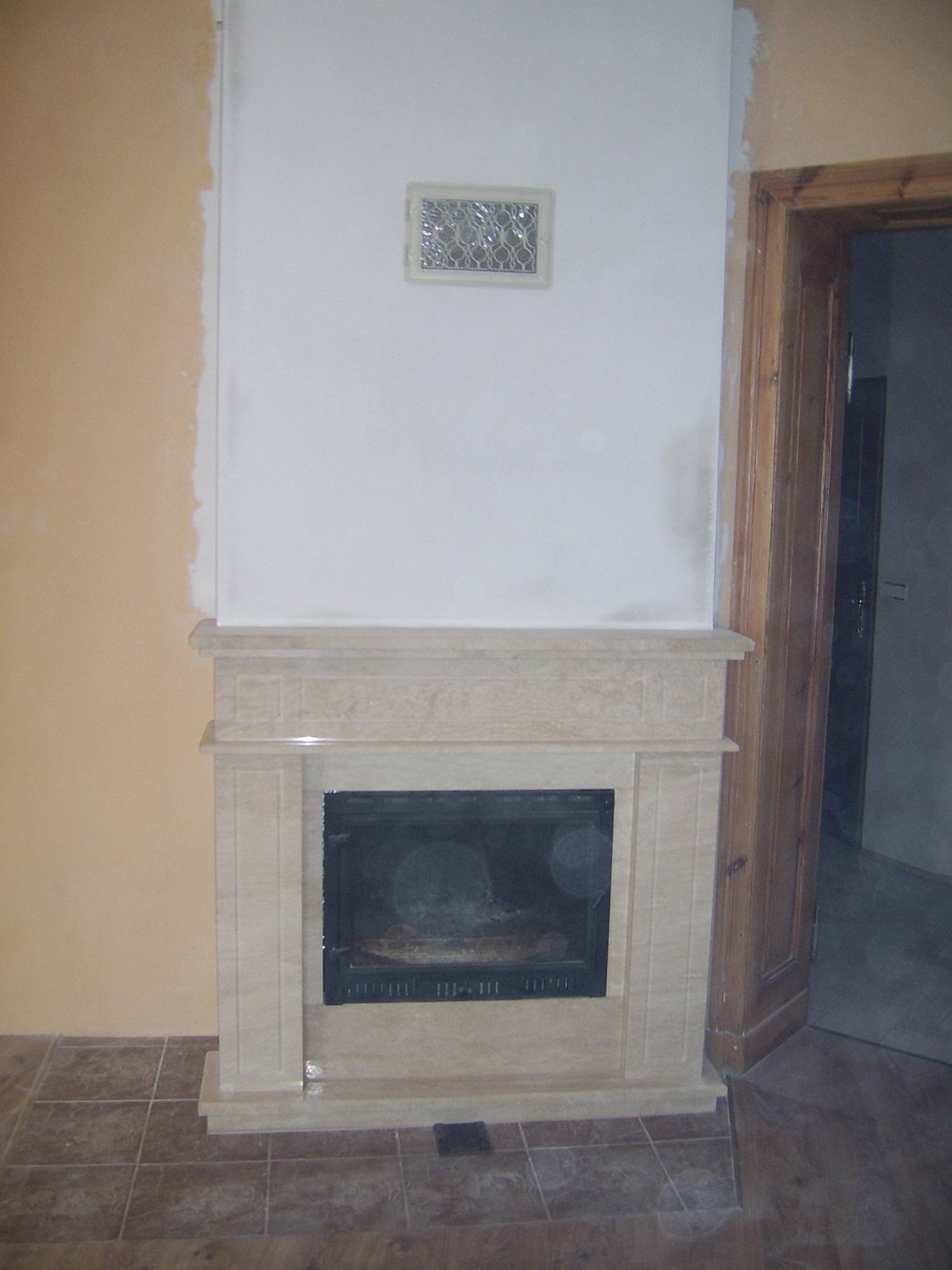 Niedokończona zabudowa kominka z beżowego kamienia z czarnym wkładem, widoczna kratka wentylacyjna na ścianie i niedomalowane fragmenty ścian.