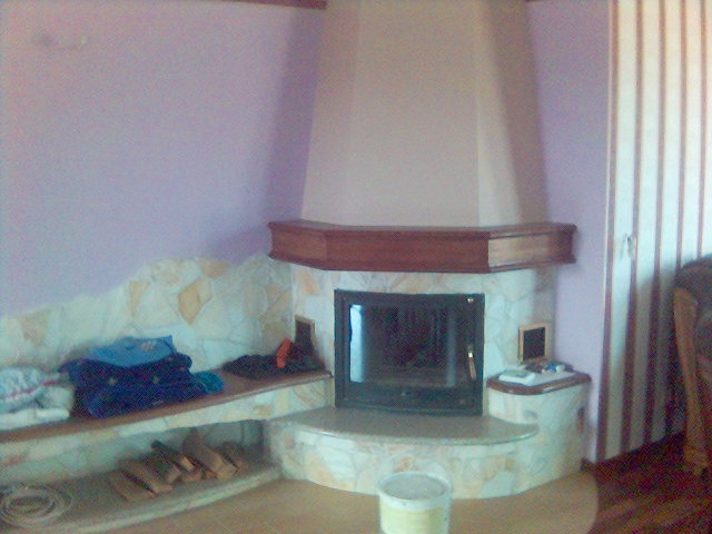 Kącik z kominkiem obłożonym kamieniem, drewniana półka nad paleniskiem, obok siedzisko z kamienia z poukładanymi ubraniami i drewnem, ściana pomalowana na liliowo.