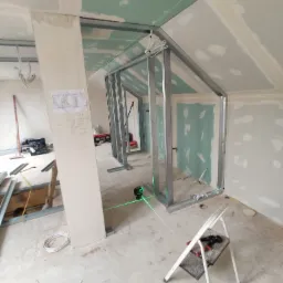 Montaż metalowej konstrukcji ścianki działowej w surowym wnętrzu poddasza, widoczny laser krzyżowy i częściowo zamontowane płyty na skosach, drabina z narzędziami w tle.