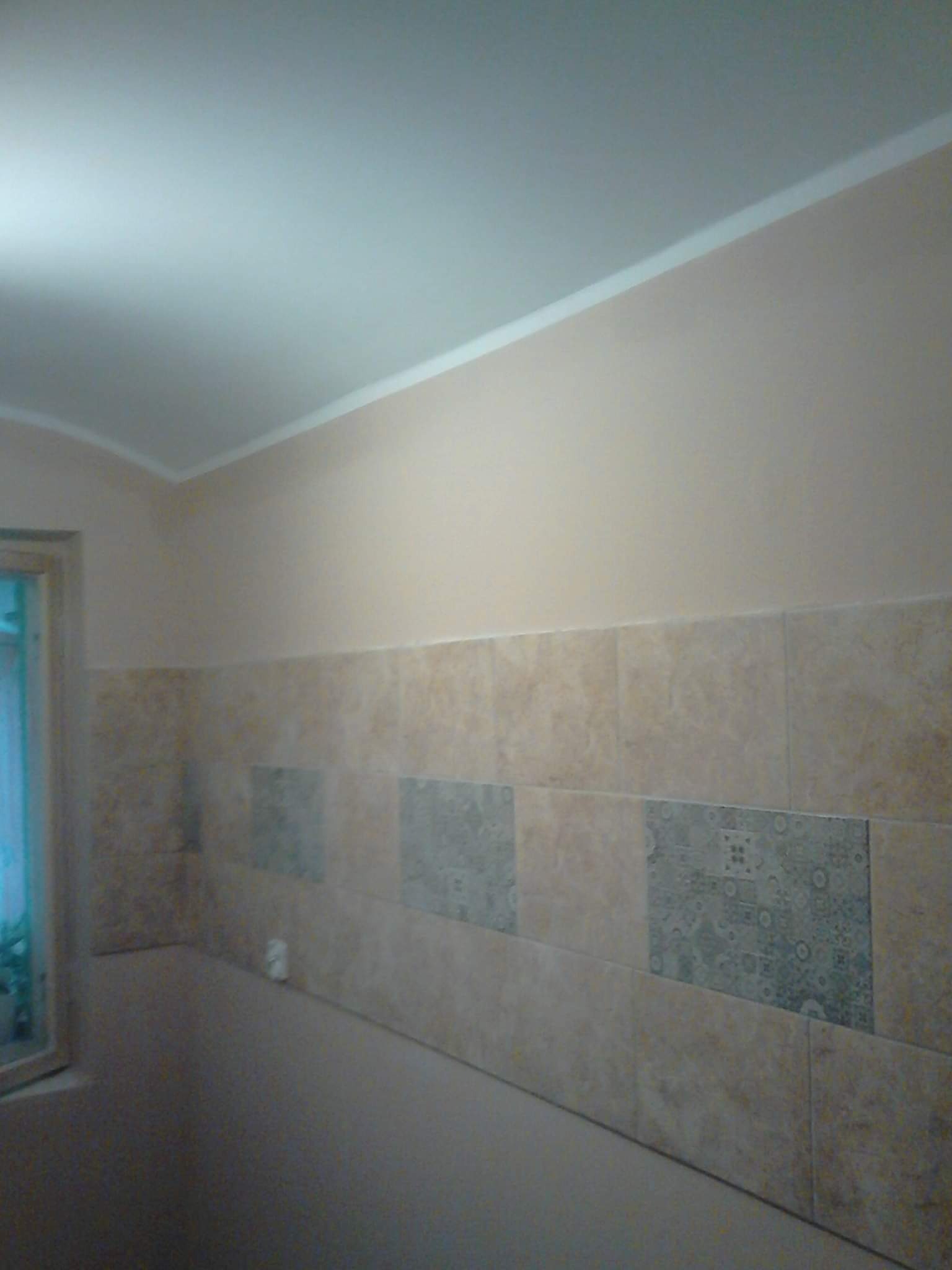 Ściana łazienki wyłożona beżowymi i szarymi płytkami ceramicznymi z wzorami, górna część ściany pomalowana na beżowo, widoczne białe listwy przy suficie i okno.