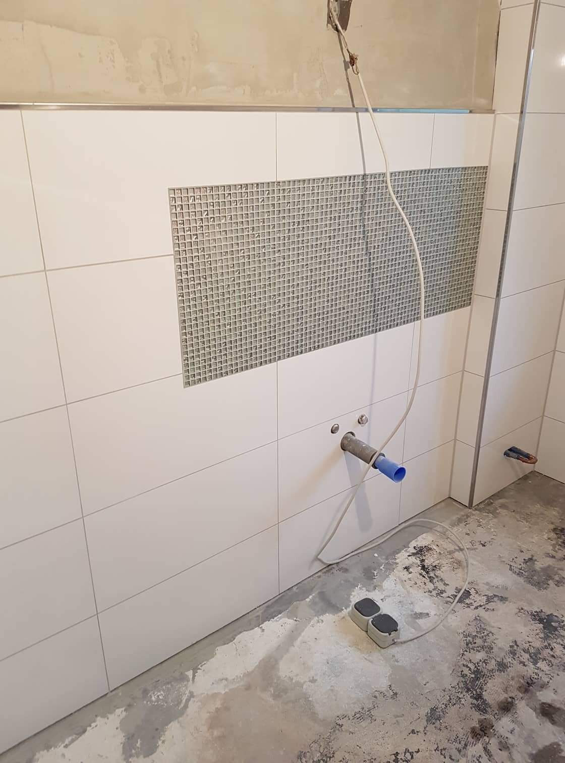 Białe płytki ścienne z szarą mozaiką w trakcie montażu w łazience, widoczne podłączenie rury z niebieskim elementem i niezabezpieczone gniazdka na podłodze.