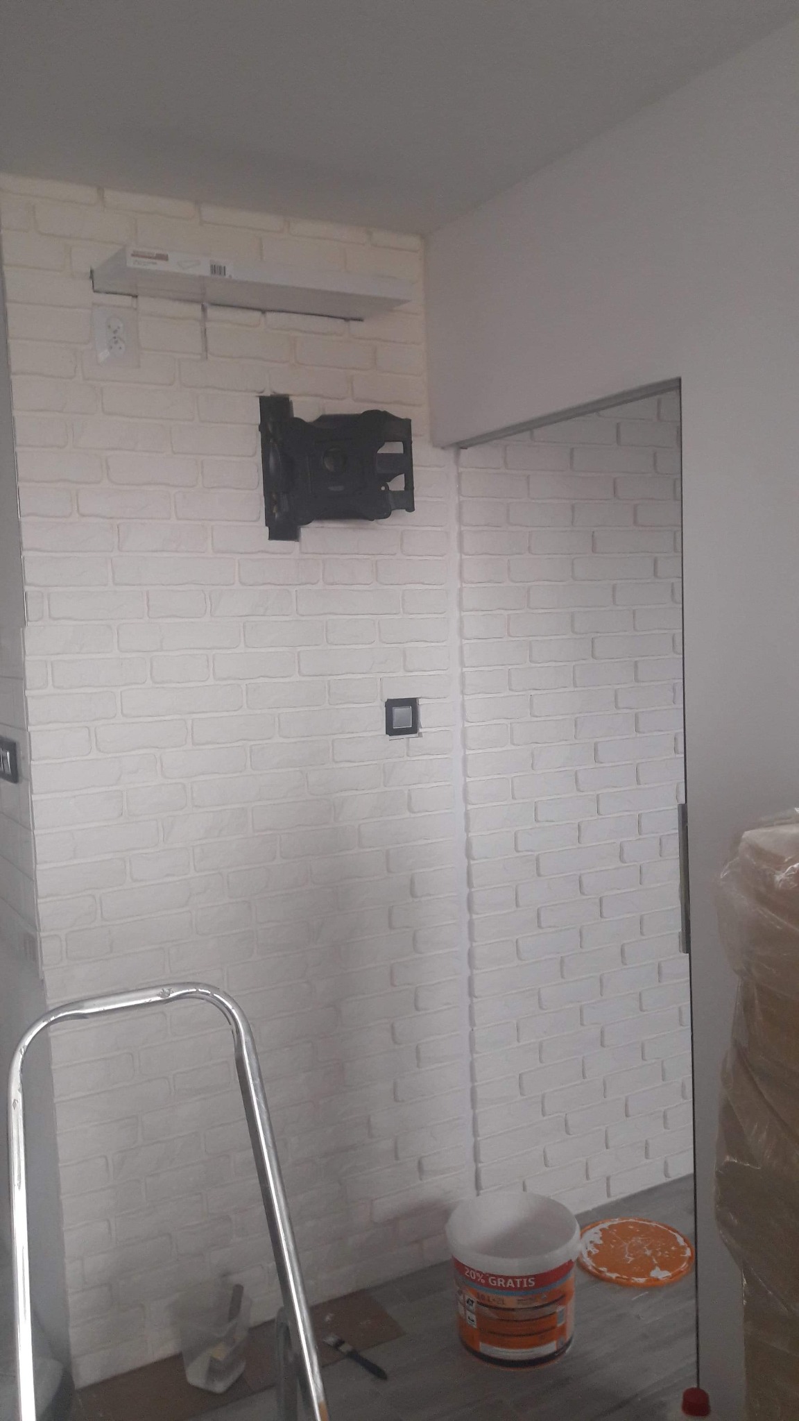 Biała ściana z cegieł, uchwyt na telewizor, puszka farby 20% gratis, drabina i widoczne ślady malowania na podłodze.