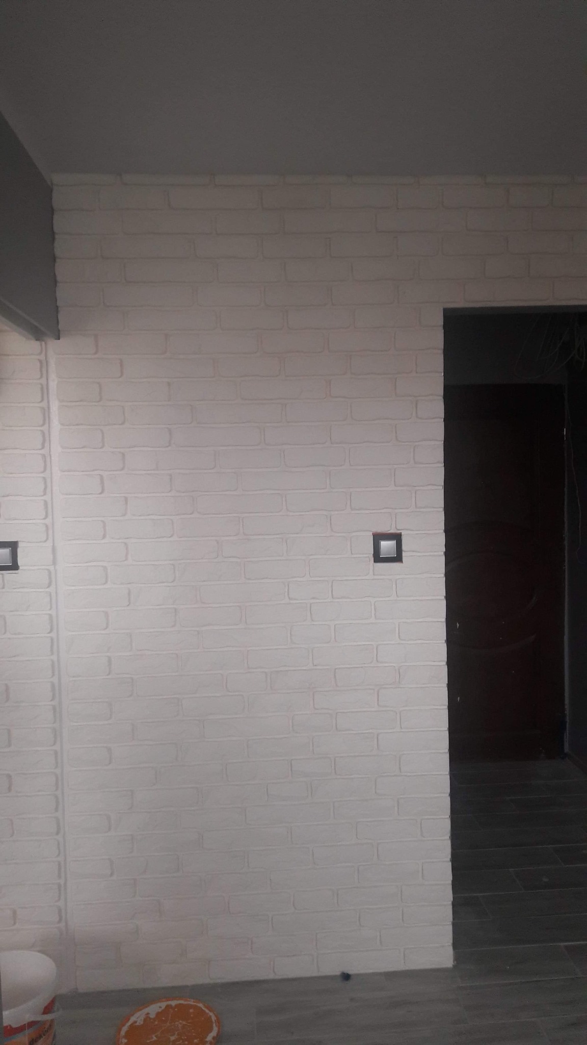 Ściana wyłożona białymi płytkami imitującymi cegłę, z zamontowanymi czarnymi włącznikami światła, widok na ciemne drzwi i fragment szarego sufitu, u dołu widoczne fragmenty akcesoriów malarskich.