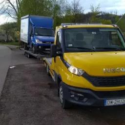 Żółta laweta Iveco z niebieskim busem na platformie, zaparkowana na poboczu, widok z przodu, tablica rejestracyjna widoczna.