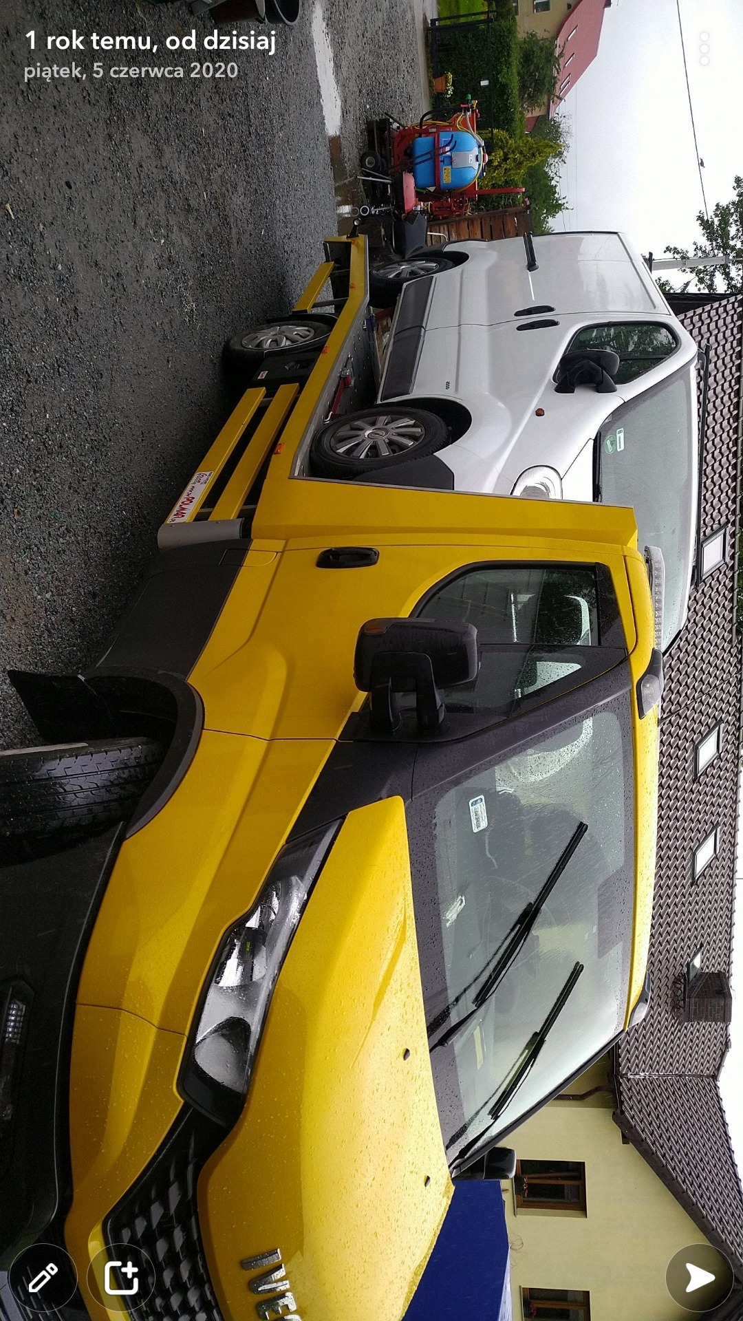 Żółta laweta z białym busem na platformie, widok z przodu na mokrej nawierzchni, w tle dom z dachówką.