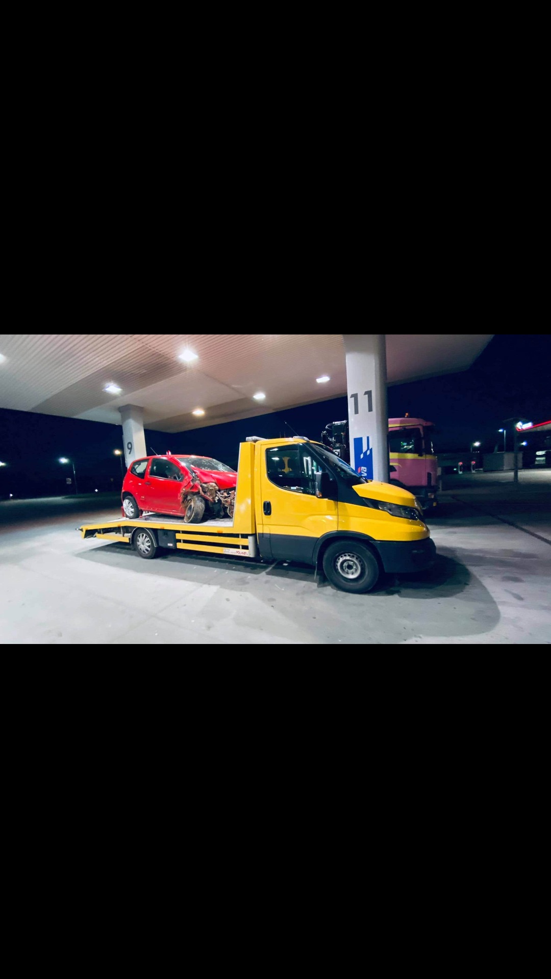 Żółta laweta z uszkodzonym czerwonym samochodem osobowym na platformie, sfotografowana na stacji benzynowej nocą.