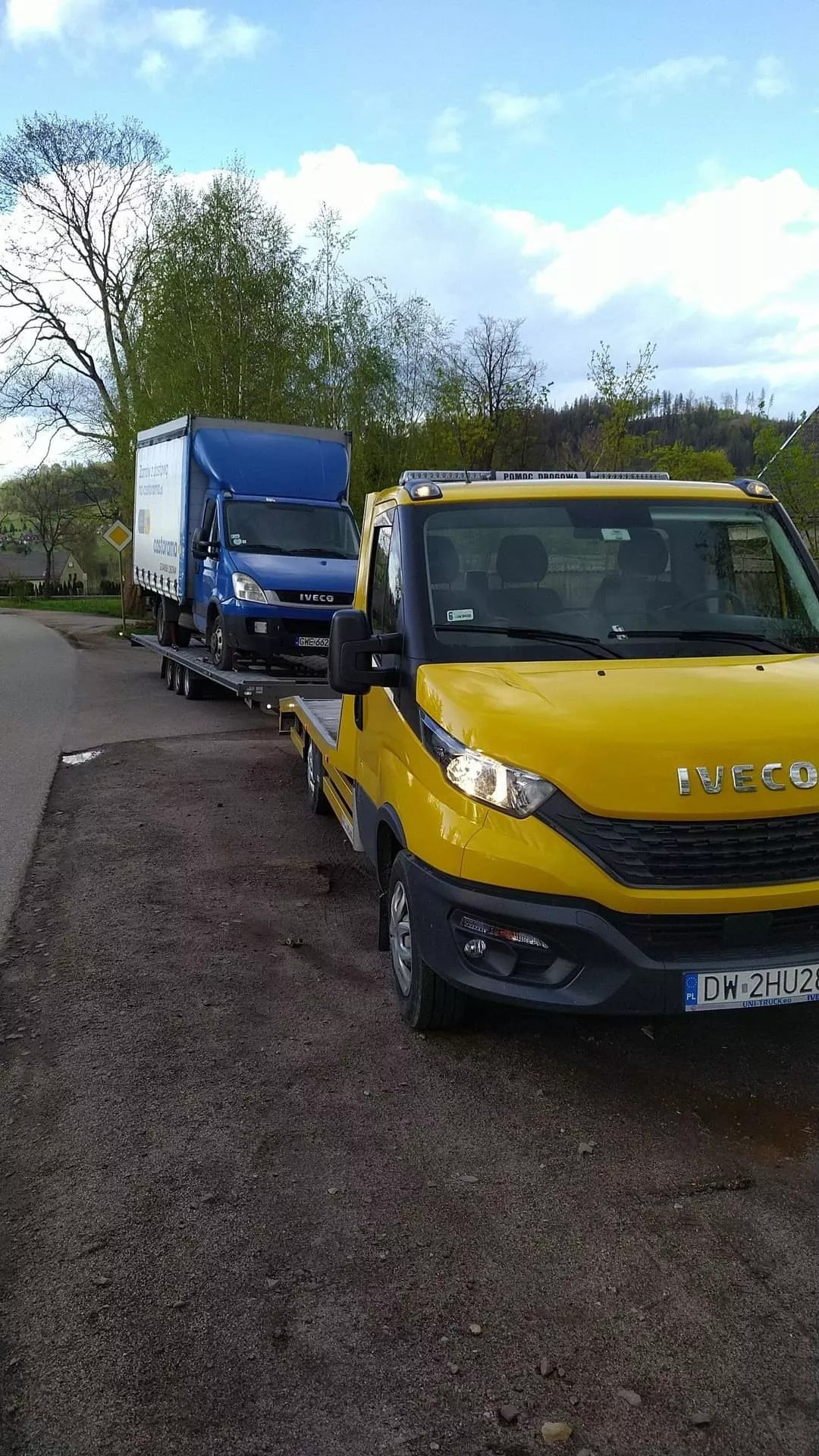 Żółta laweta Iveco z niebieskim busem na platformie, zaparkowana na poboczu, widok z przodu, tablica rejestracyjna widoczna.