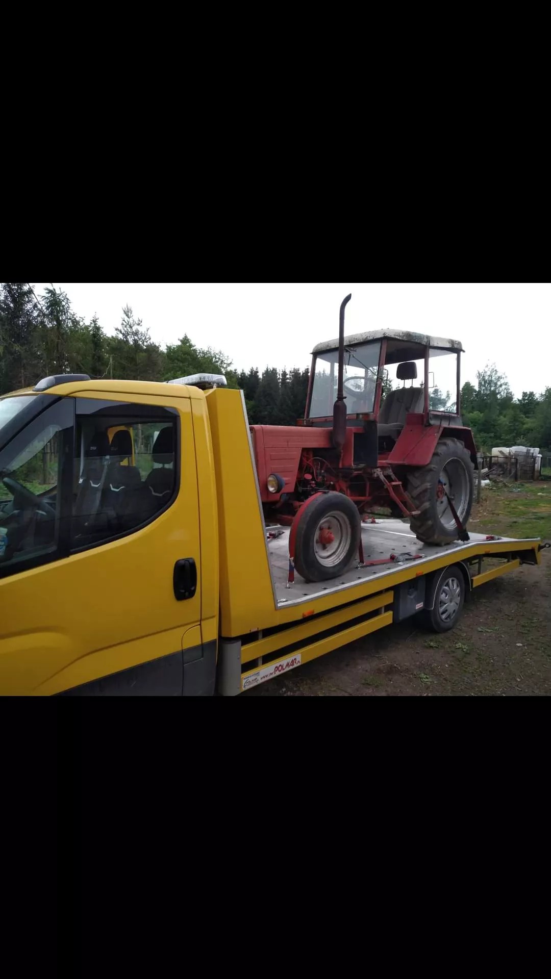 Żółta laweta z czerwonym traktorem zabezpieczonym pasami transportowymi na platformie, widok z boku.