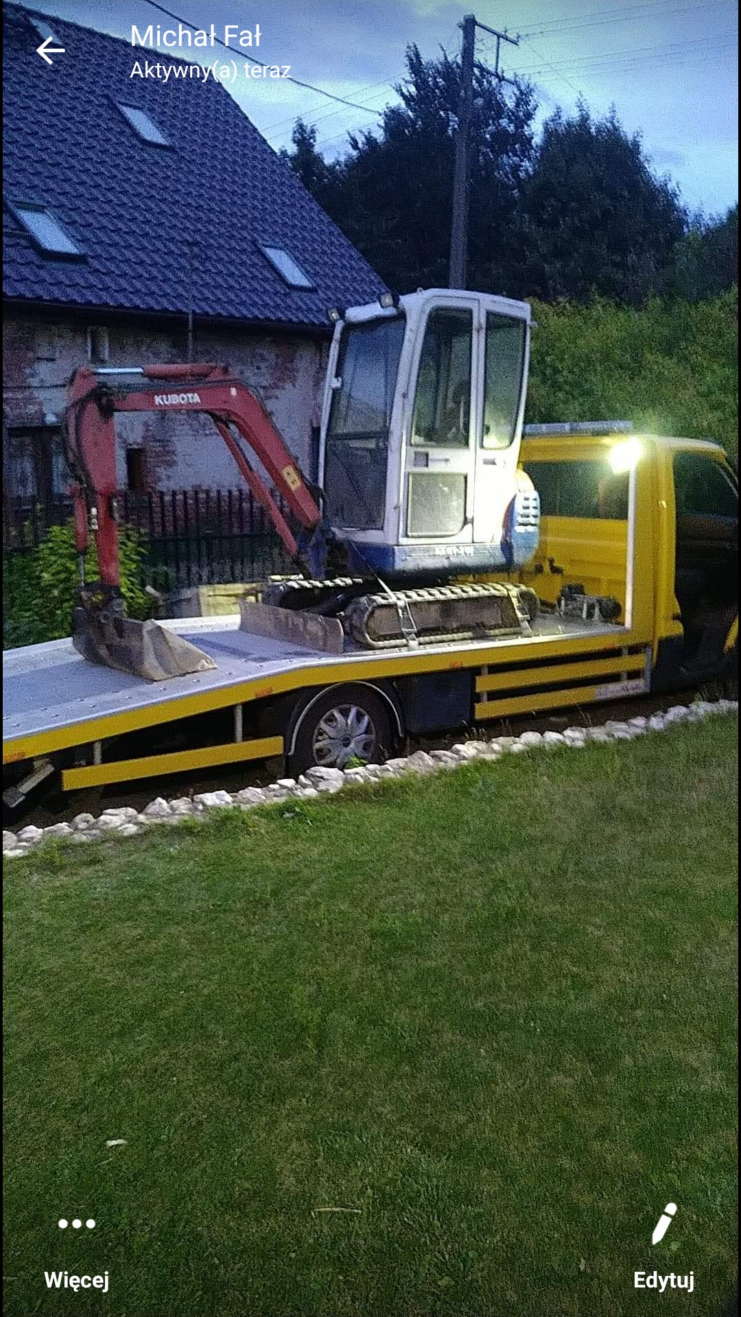 Żółta laweta z mini koparką Kubota na platformie transportowej, zaparkowana na trawniku przed domem.