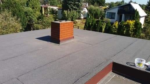 Widok z góry na płaski dach pokryty papą z wystającym ceglanym kominem. W tle widać domy i zieleń. Na dachu, w prawym dolnym rogu, stoi wiaderko z narzędziami.