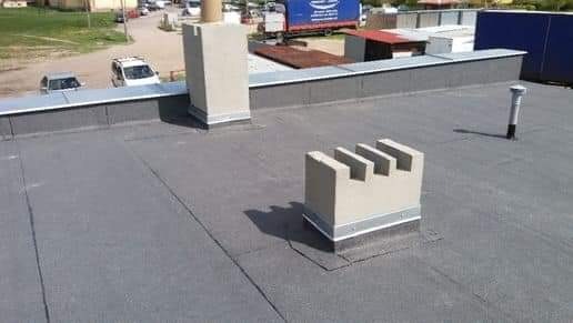 Wykończony, płaski dach pokryty papą z dwoma kominami, jeden z trzema wlotami wentylacyjnymi, drugi wysoki, kwadratowy, w tle budynki i samochody.