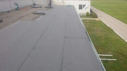 Szary, płaski dach budynku komercyjnego z widocznymi łączeniami papy, fragmentem instalacji na krawędzi i budynkiem w tle.