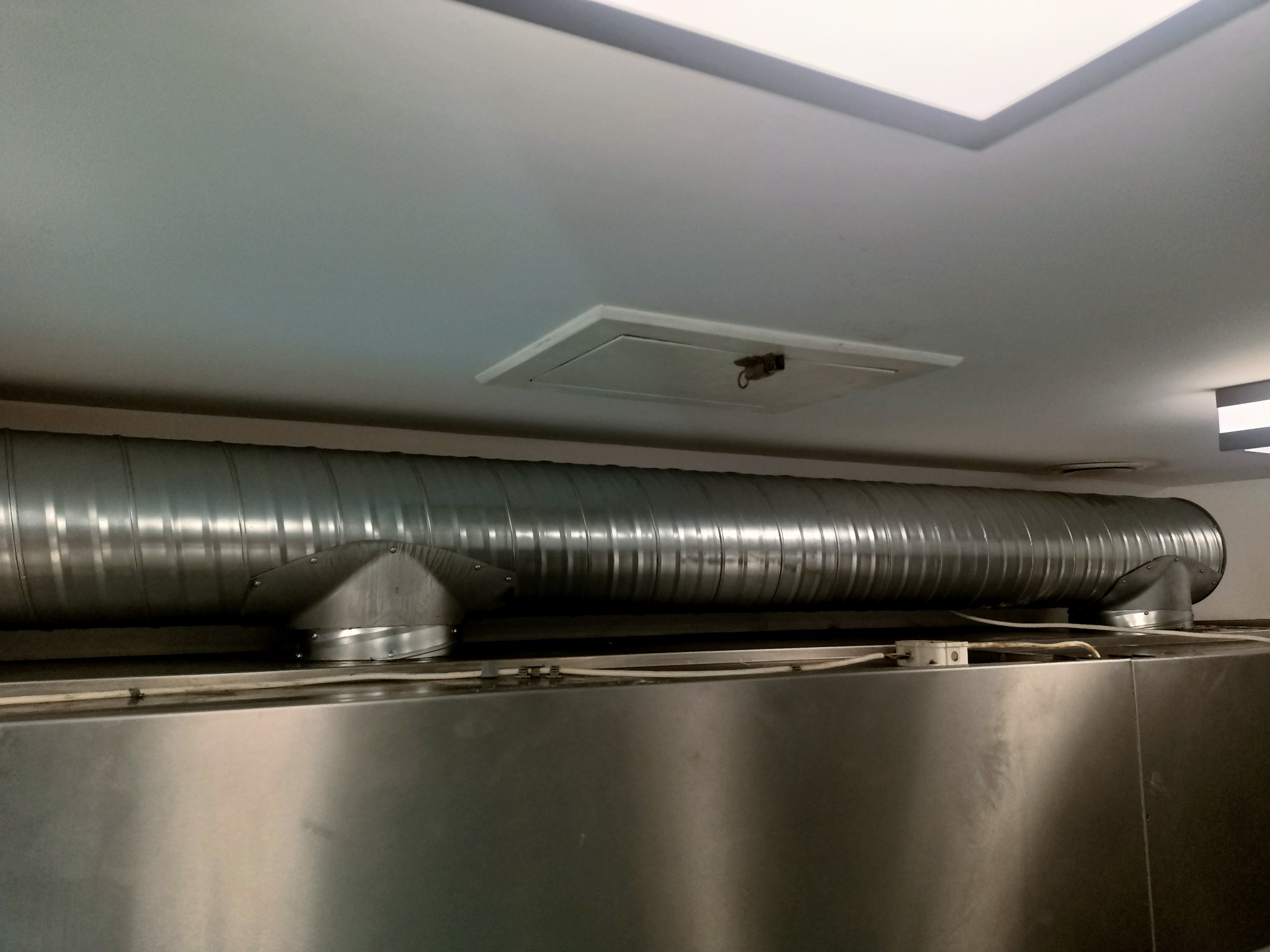 Sztywny, spiralny przewód wentylacyjny z metalu, zamontowany pod sufitem w pomieszczeniu z metalowymi powierzchniami. Widoczna klapa rewizyjna w suficie.