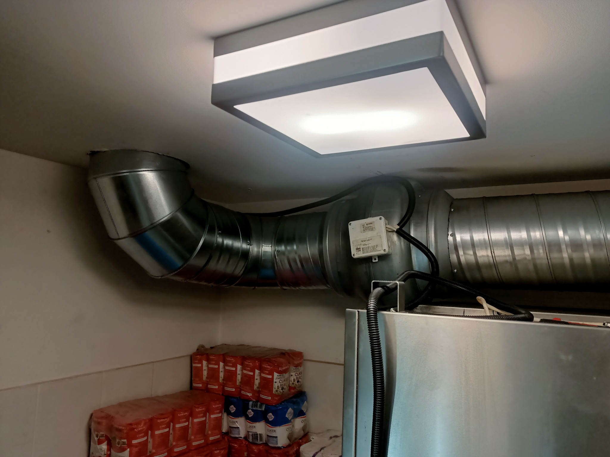 Metalowy kanał wentylacyjny, z widocznym czujnikiem, biegnący pod sufitem obok kwadratowej lampy sufitowej, z zapasem cukru w tle.