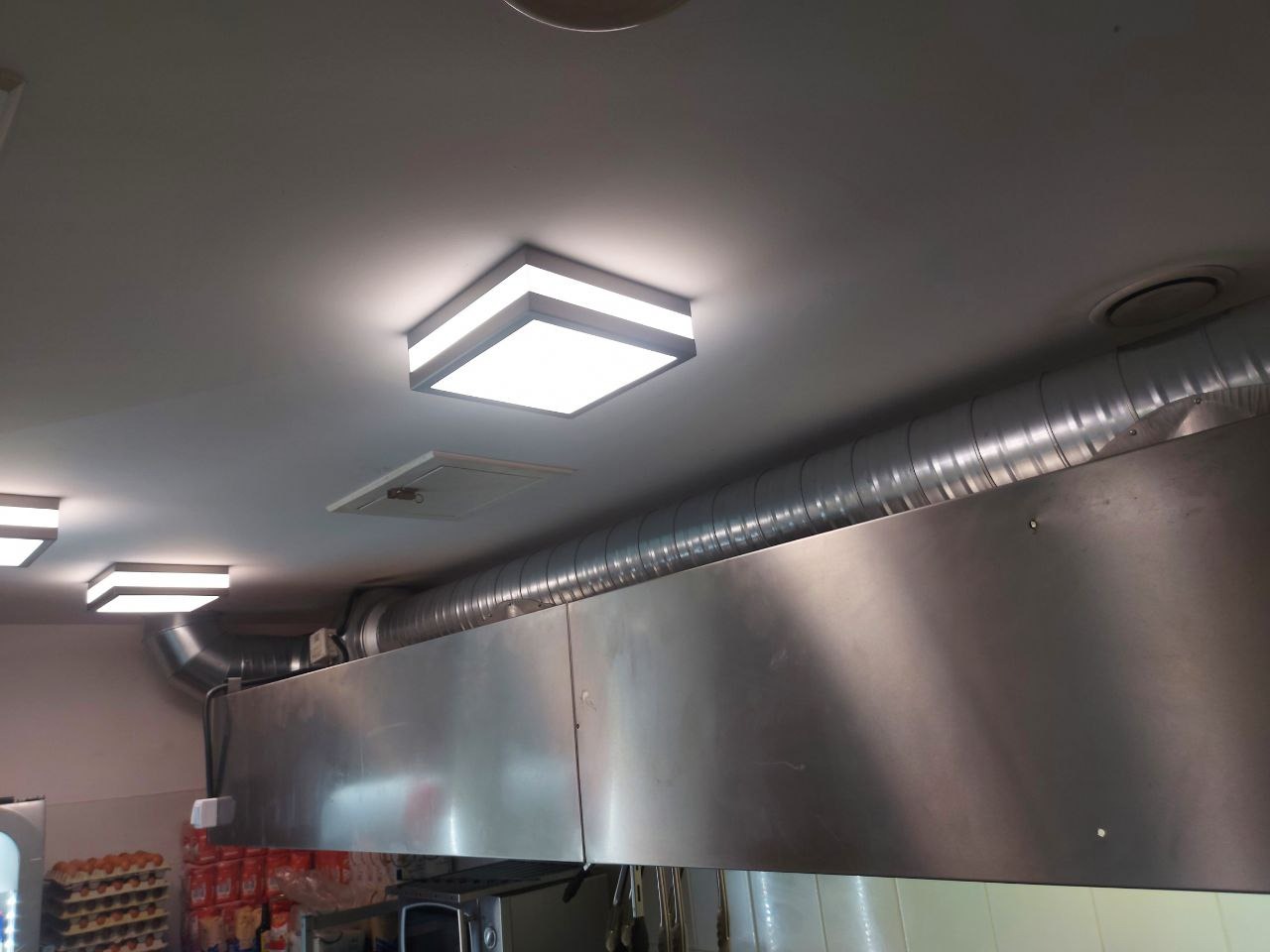 Widok na sufit kuchni komercyjnej z zainstalowaną, metalową instalacją wentylacyjną z rurami spiro, oświetleniem LED w kwadratowych oprawach i kratką rewizyjną.