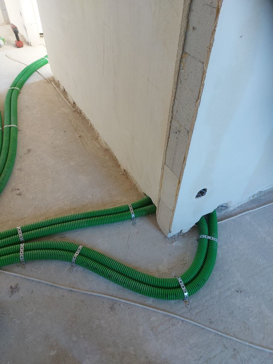 Zielone, karbowane rury wentylacyjne przymocowane metalowymi obejmami do ściany w surowym stanie, widoczny otwór w ścianie i przewód elektryczny na podłodze.