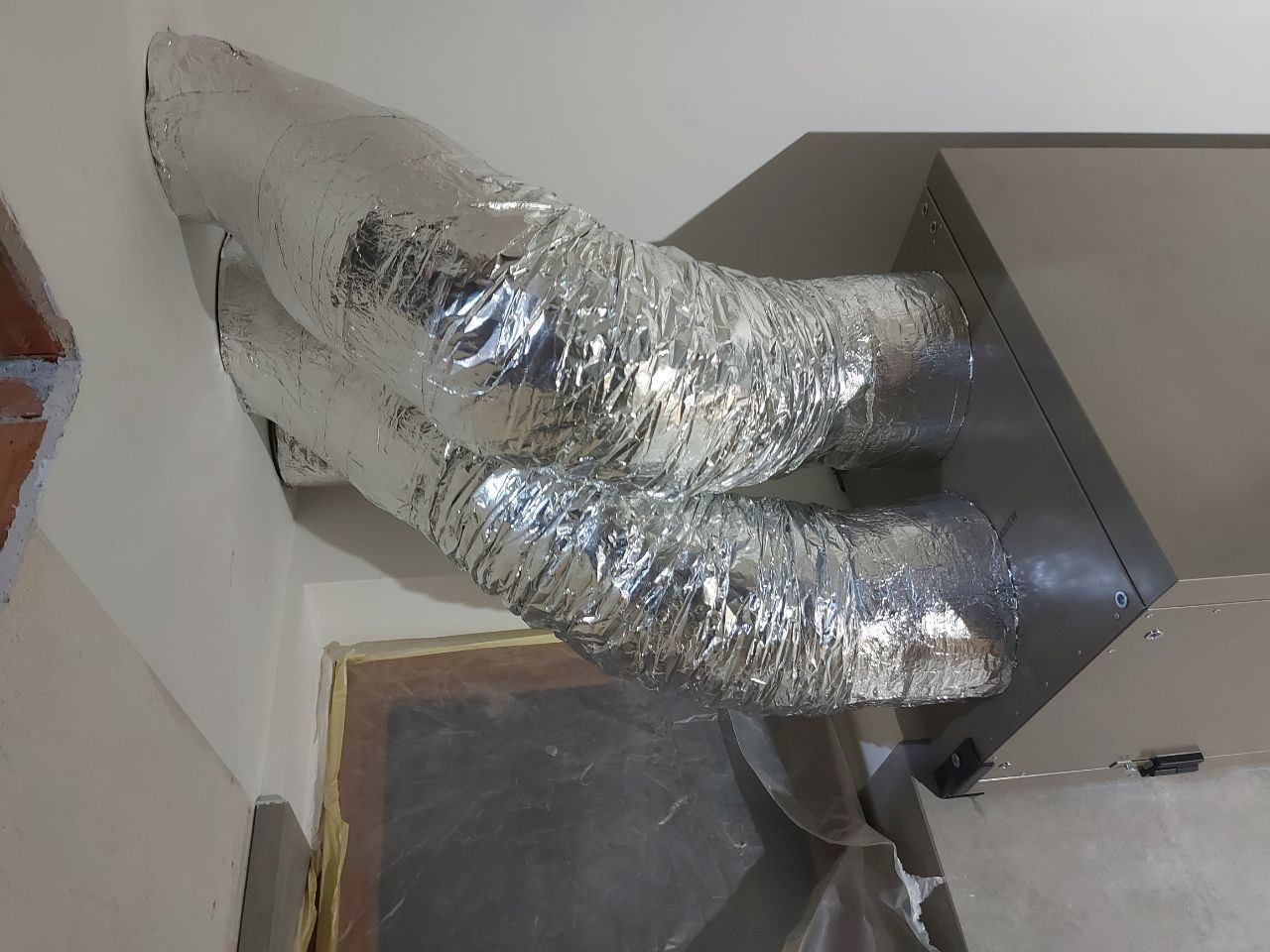 Dwa srebrne, izolowane przewody wentylacyjne podłączone do szarej jednostki centralnej systemu wentylacji, widoczne elementy zabezpieczenia okna folią malarską.