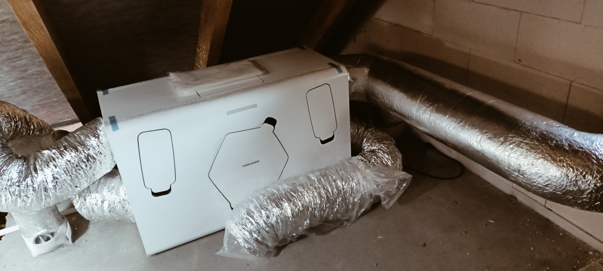 Białe opakowanie rekuperatora marki Thessla Green leżące na podłodze w pobliżu elastycznych przewodów wentylacyjnych w pomieszczeniu o surowym wykończeniu.