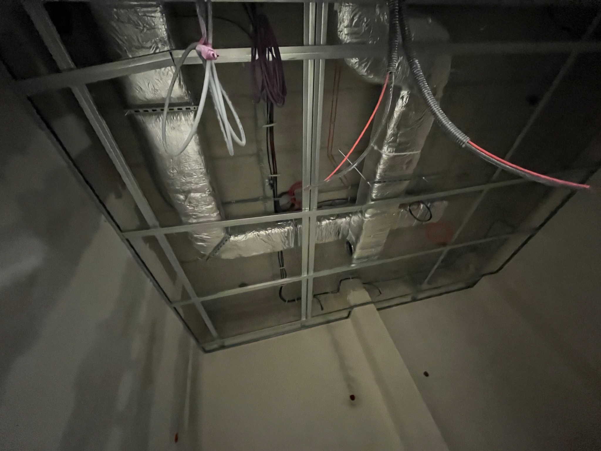 Widok z dołu na sufit podwieszany w trakcie montażu instalacji wentylacyjnej, widoczne przewody wentylacyjne w izolacji aluminiowej, kable elektryczne i rurki, wszystko zamocowane do metalowej...