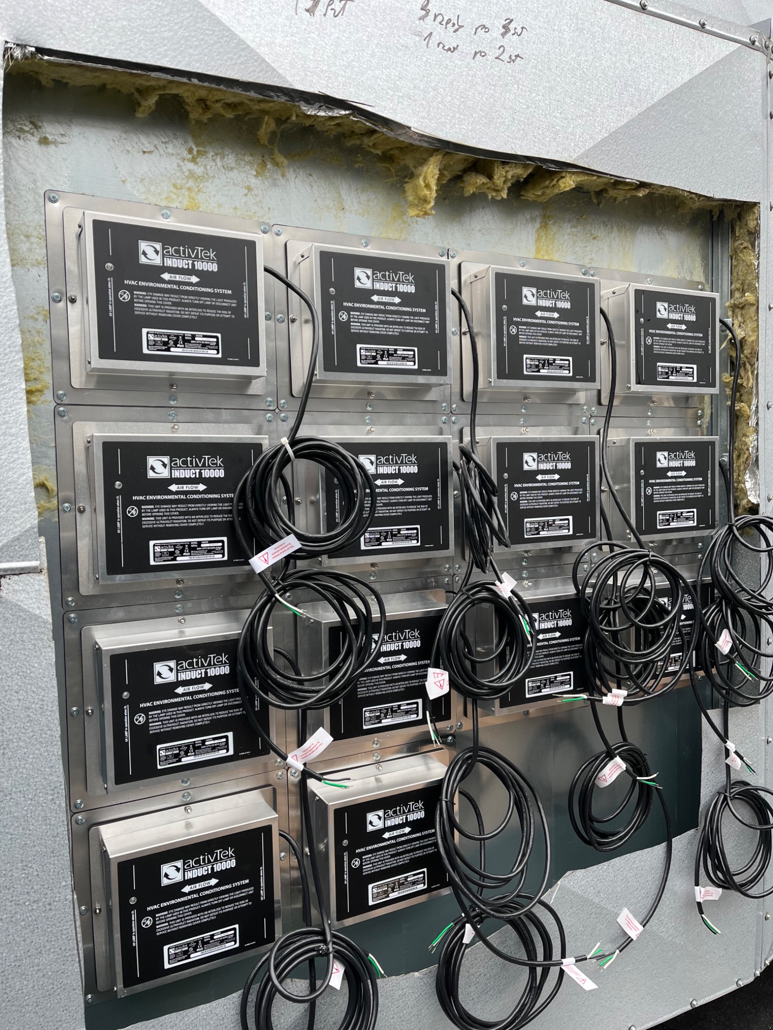 Rząd identycznych modułów ActivTek Induct 10000, zamontowanych wewnątrz kanału wentylacyjnego, z widoczną izolacją z wełny mineralnej w górnej części. Z każdego modułu wychodzą zwinięte kable...