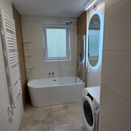 Kompleksowe wykończenia wnętrz. Jacek Sacharczuk - Nowoczesna łazienka z wanną, grzejnikiem drabinkowym, pralką i podświetlanym lustrem. Jasne płytki, szklana ścianka prysznicowa i okno nad wanną.