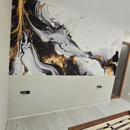 Kompleksowe wykończenia wnętrz. Jacek Sacharczuk - Pokój w trakcie dekoracji z abstrakcyjną tapetą w odcieniach bieli, czerni i złota na górnej części ściany. Widoczne gniazdka elektryczne i fragment drewnianego łóżka.