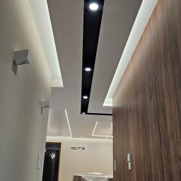 Kompleksowe wykończenia wnętrz. Jacek Sacharczuk - Nowoczesny sufit podwieszany z oświetleniem LED i czarnymi reflektorami, widok z dołu, drewniane drzwi po prawej, wnętrze w trakcie wykończenia.