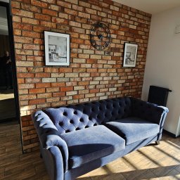 Kompleksowe wykończenia wnętrz. Jacek Sacharczuk - Błękitna sofa chesterfield na tle ceglanej ściany z zegarem i obrazami. Parkiet w jodełkę i oświetlenie punktowe tworzą stylowe wnętrze.