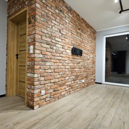 Kompleksowe wykończenia wnętrz. Jacek Sacharczuk - Wnętrze po remoncie: ceglana ściana z zamontowanym panelem sterowania, drewniane drzwi i podłoga z płytek imitujących drewno. Widok na sąsiednie pomieszczenie.