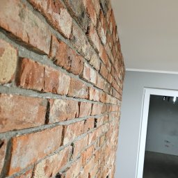 Kompleksowe wykończenia wnętrz. Jacek Sacharczuk - Dekoracyjna ściana z cegły w odcieniach czerwieni i brązu, z widocznym uchwytem TV,  w nowoczesnym wnętrzu z białymi drzwiami i szarą ścianą. Nietypowy kadr.