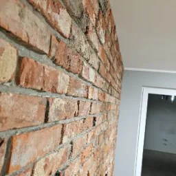 Dekoracyjna ściana z cegły w odcieniach czerwieni i brązu, z widocznym uchwytem TV,  w nowoczesnym wnętrzu z białymi drzwiami i szarą ścianą. Nietypowy kadr.