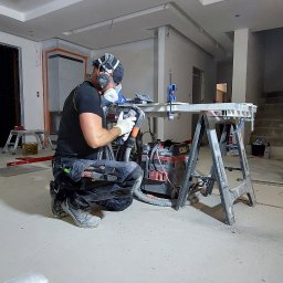 Kompleksowe wykończenia wnętrz. Jacek Sacharczuk - Pracownik w masce szlifuje element w surowym wnętrzu. Widoczny pył, narzędzia i prowizoryczny stół warsztatowy. W tle fragmenty ścian i sufitu.