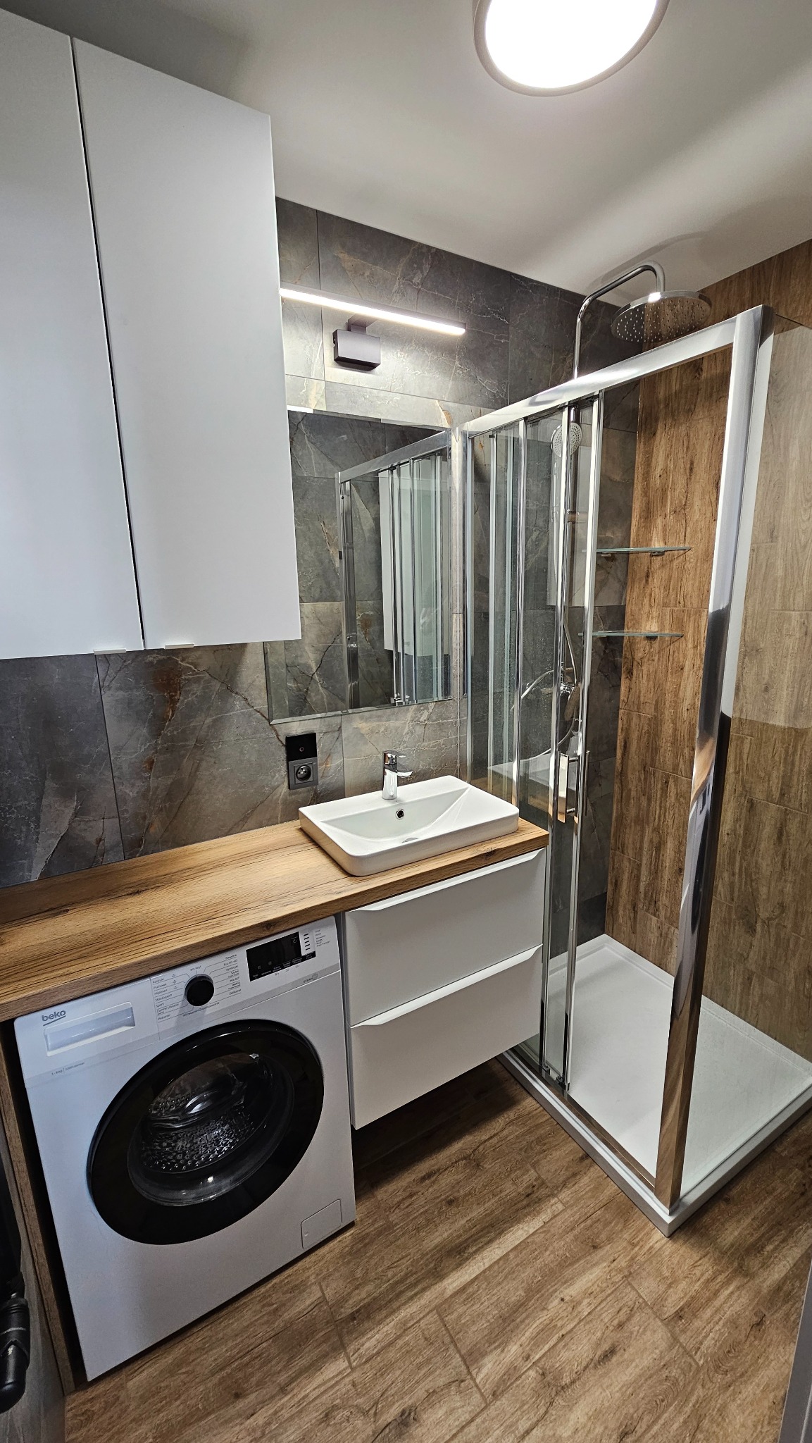 Nowoczesna łazienka z kabiną prysznicową typu walk-in, umywalką nablatową i pralką pod blatem z drewnianym wykończeniem. Aranżacja w szaro-brązowej kolorystyce.