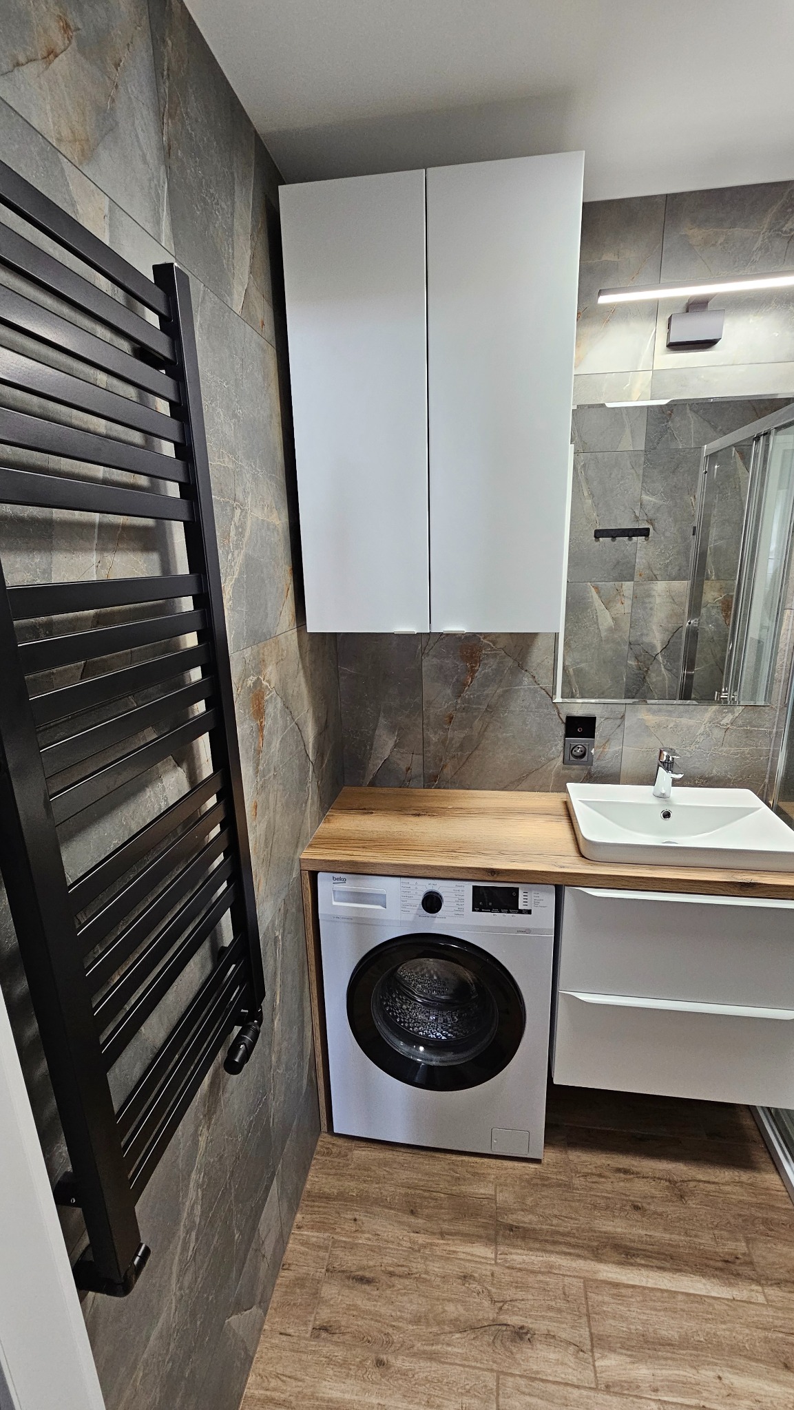 Wykończona łazienka w Szczecinie z pralką pod blatem, umywalką i grzejnikiem drabinkowym na tle szarej, kamiennej mozaiki ściennej i drewnopodobnych płytek podłogowych.
