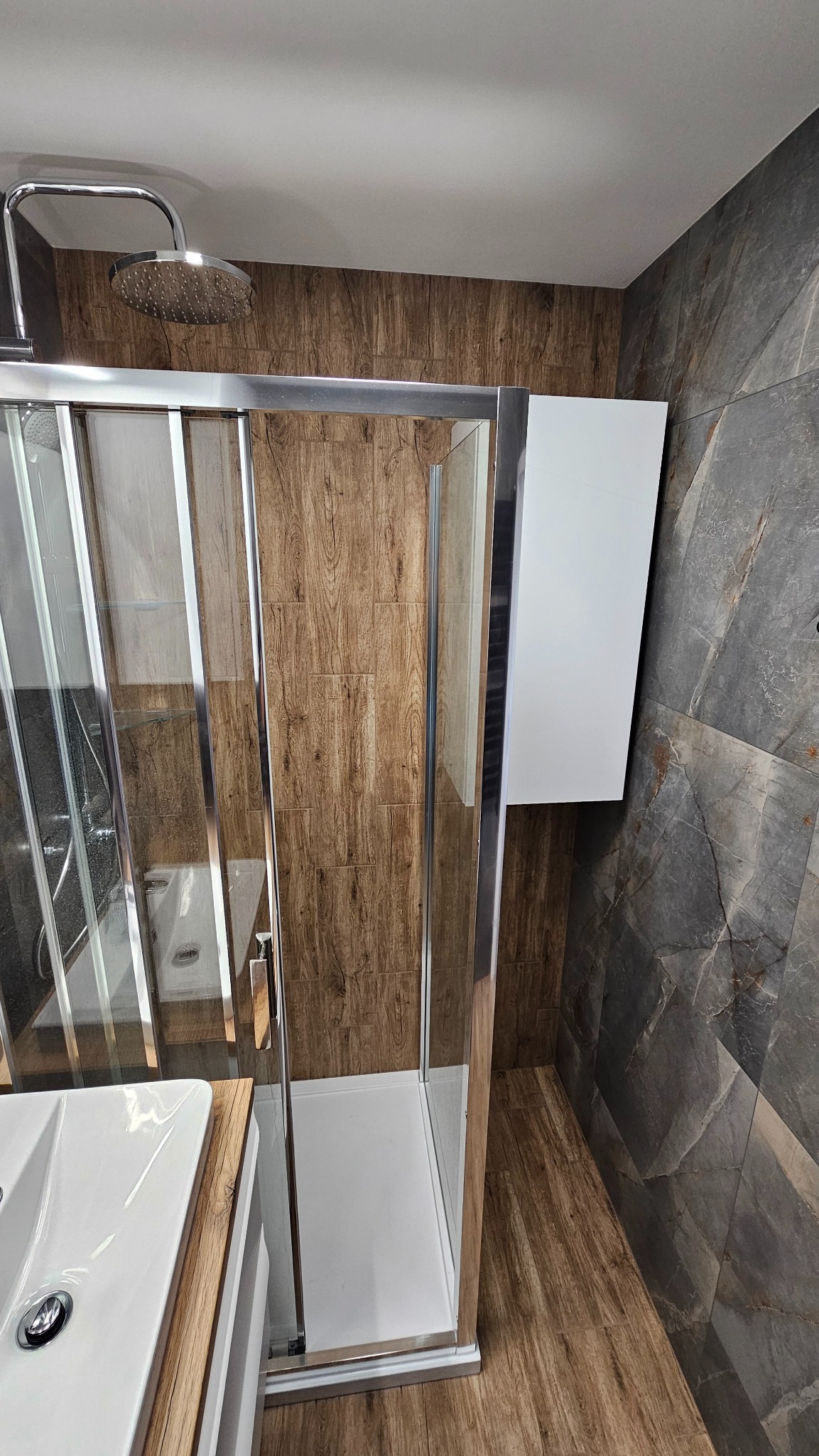 Nowoczesna łazienka z kabiną prysznicową ze srebrnymi ramami, drewnopodobnymi i marmurowymi płytkami oraz białą szafką wiszącą. Widok na umywalkę.