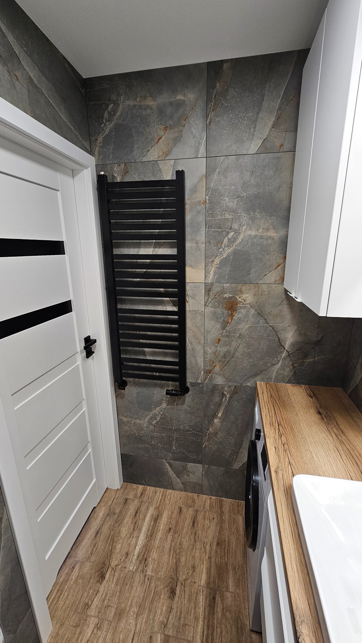 Wykończona łazienka z szarymi płytkami imitującymi marmur, drewnopodobną podłogą, białymi szafkami i drzwiami z czarnymi akcentami oraz czarnym grzejnikiem.