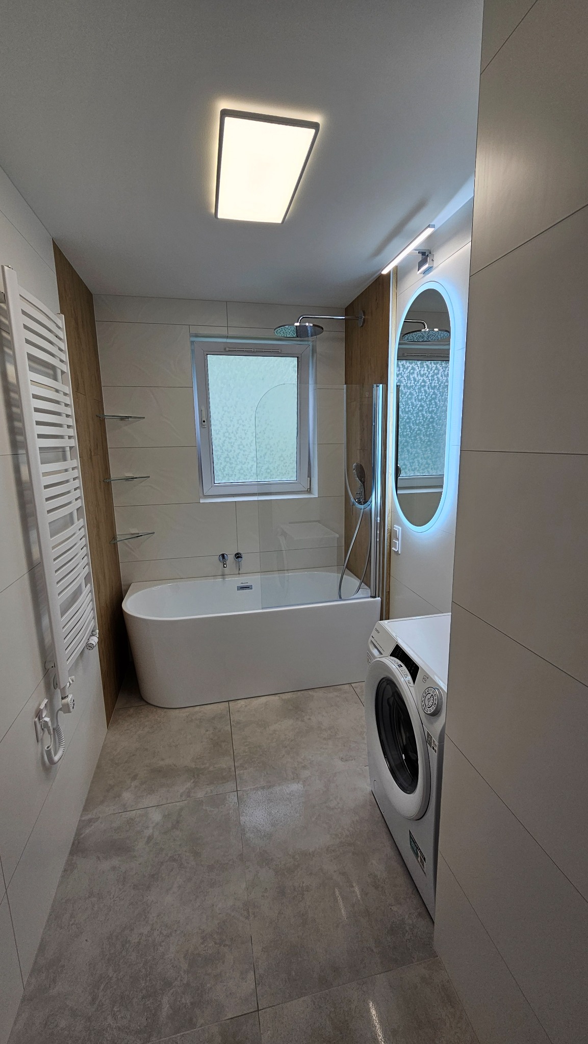 Nowoczesna łazienka z wanną, grzejnikiem drabinkowym, pralką i podświetlanym lustrem. Jasne płytki, szklana ścianka prysznicowa i okno nad wanną.