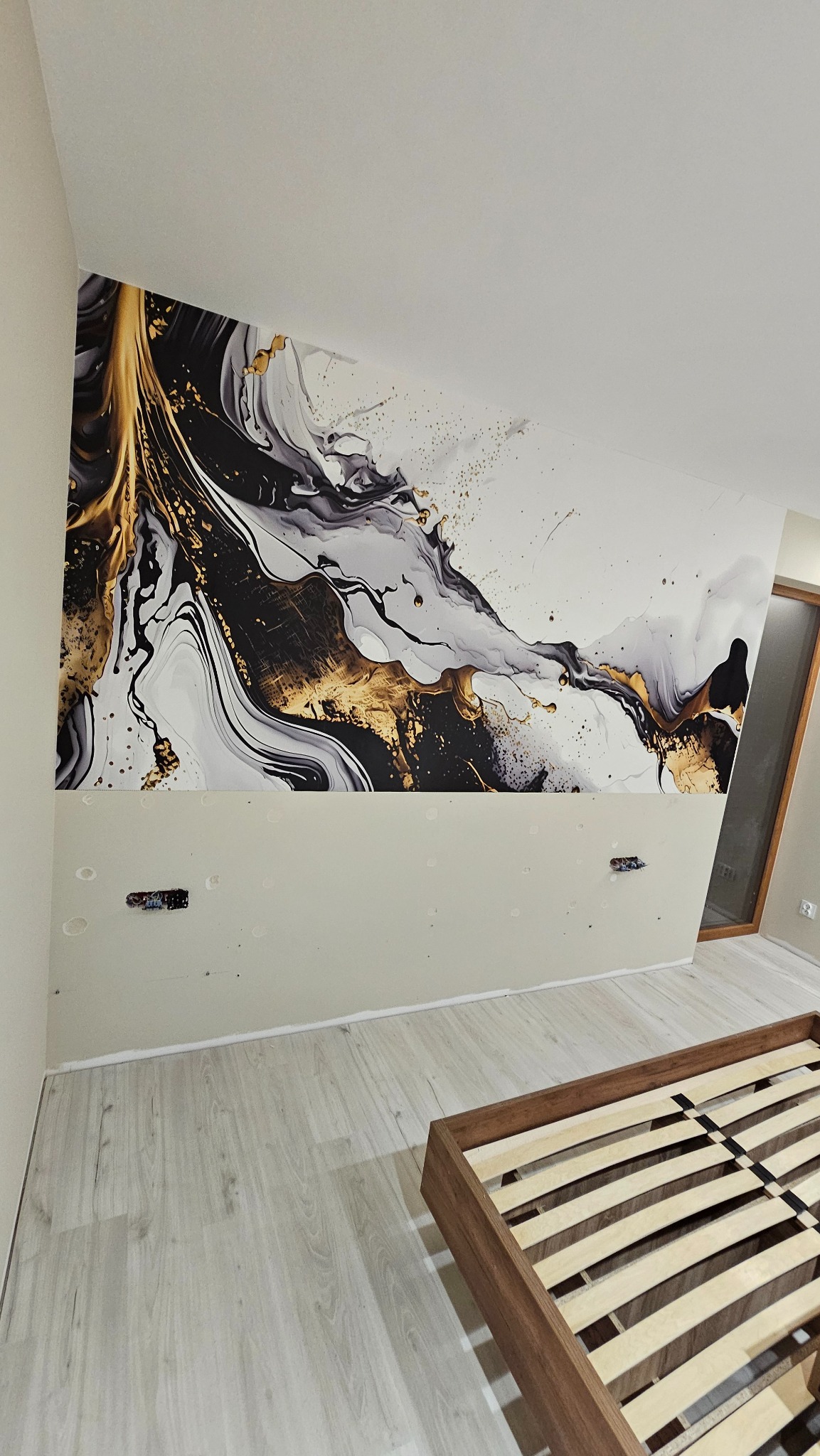 Pokój w trakcie dekoracji z abstrakcyjną tapetą w odcieniach bieli, czerni i złota na górnej części ściany. Widoczne gniazdka elektryczne i fragment drewnianego łóżka.