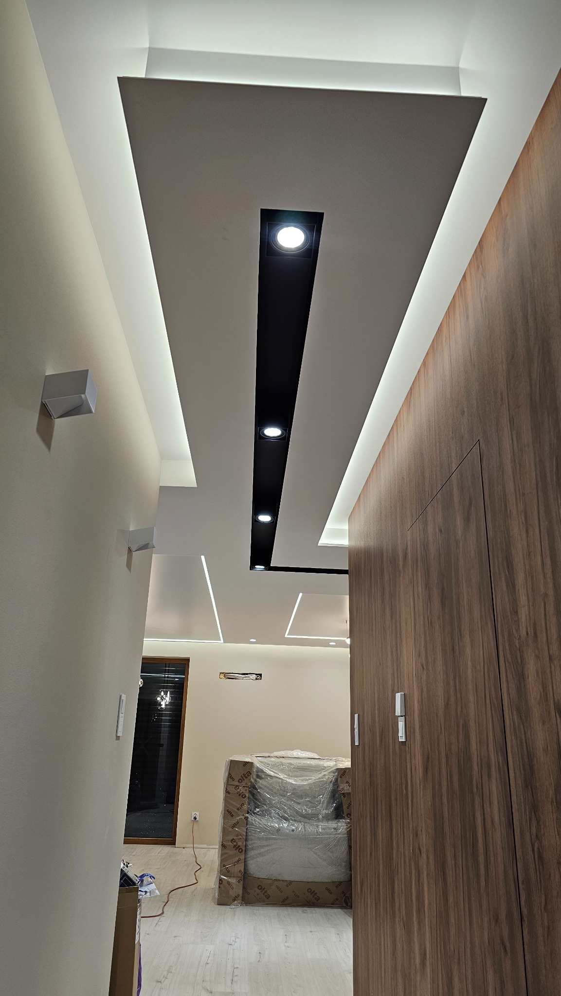 Nowoczesny sufit podwieszany z oświetleniem LED i czarnymi reflektorami, widok z dołu, drewniane drzwi po prawej, wnętrze w trakcie wykończenia.