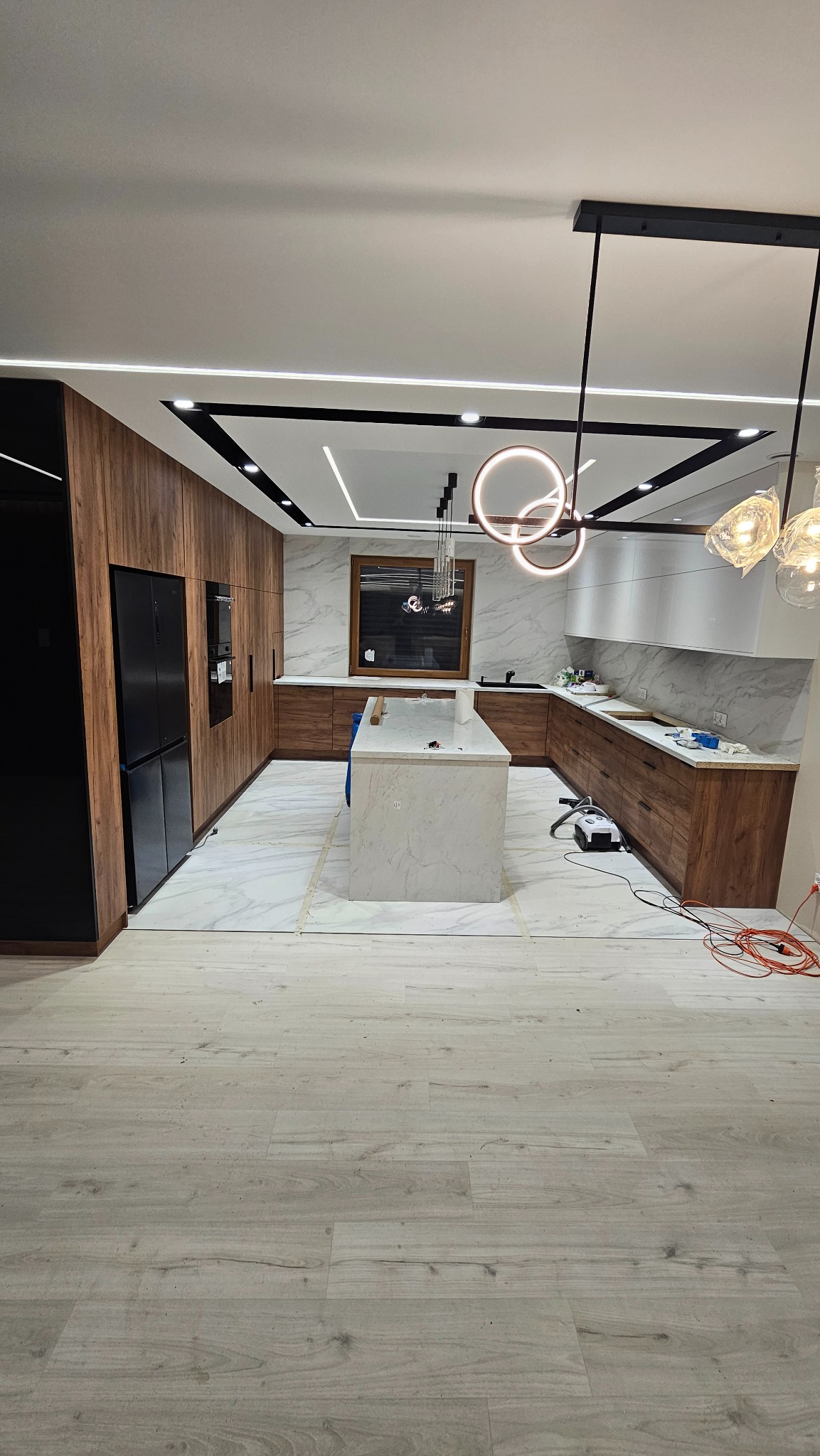 Nowoczesna kuchnia z wyspą, drewnianymi szafkami i marmurowymi płytkami. Widoczne oświetlenie sufitowe i podłoga wyłożona panelami. Sprzęt czyszczący na podłodze.