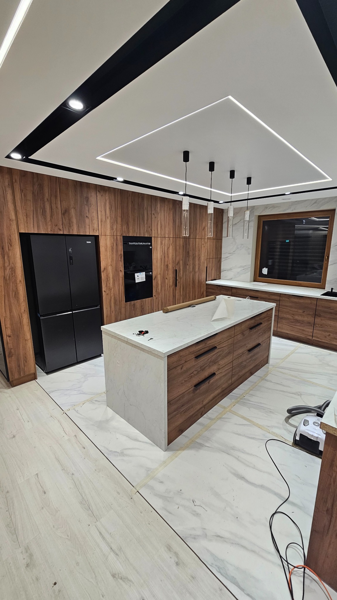 Nowoczesna kuchnia z wyspą w trakcie wykańczania: drewniane szafki, marmurowe blaty, oświetlenie LED na suficie. Widoczne niedokończone podłogi i sprzęt AGD.