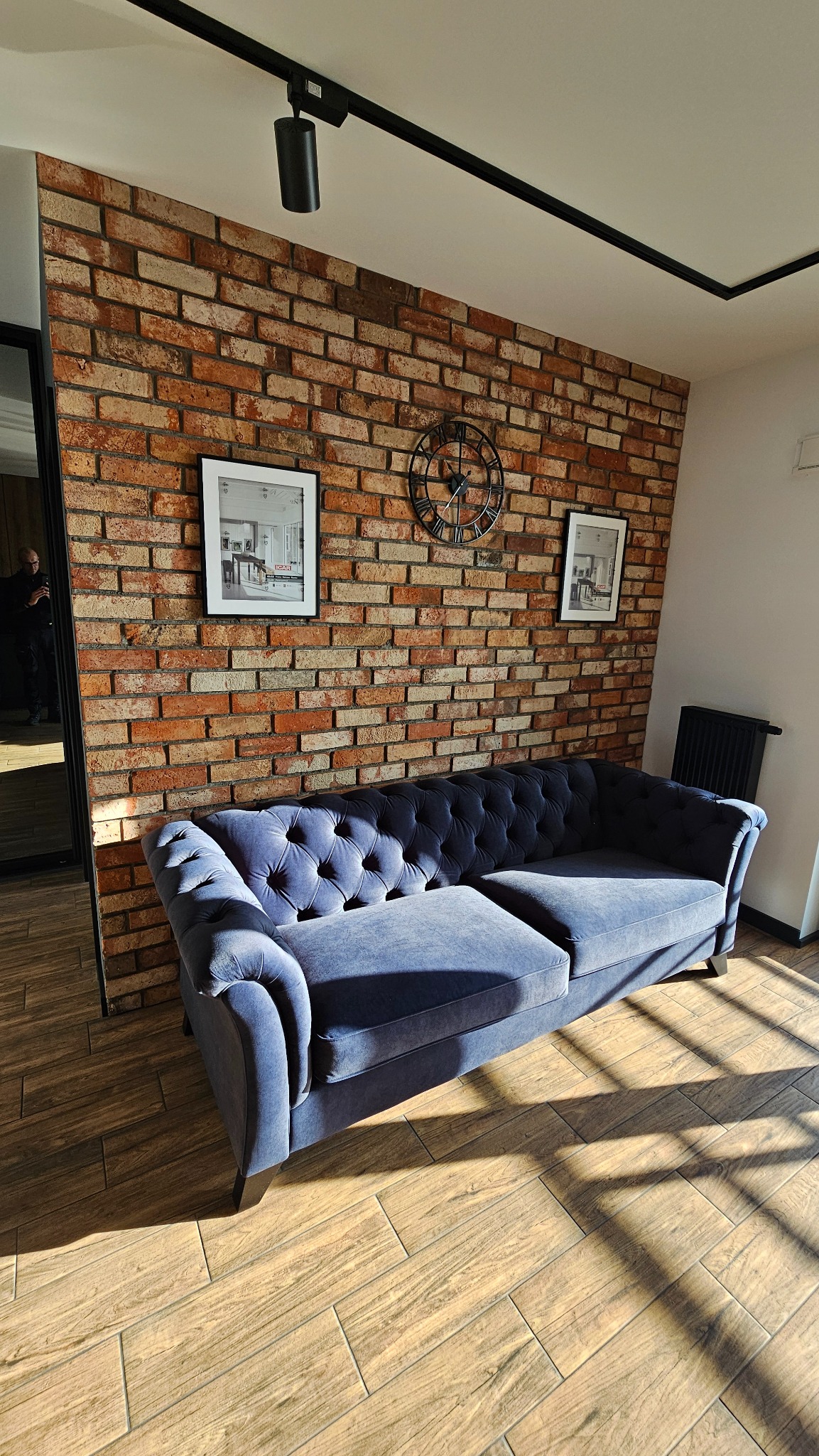 Błękitna sofa chesterfield na tle ceglanej ściany z zegarem i obrazami. Parkiet w jodełkę i oświetlenie punktowe tworzą stylowe wnętrze.