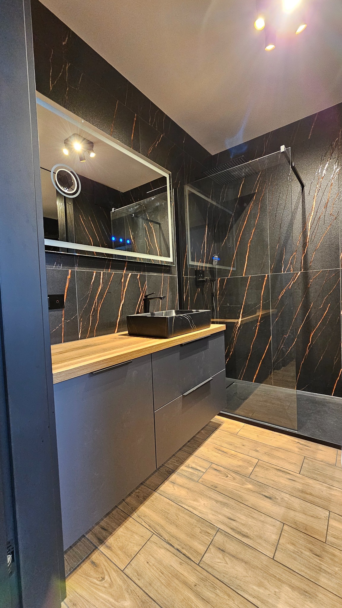 Nowoczesna łazienka z czarnymi płytkami imitującymi marmur z miedzianymi żyłami, drewnopodobną podłogą i szarą szafką pod umywalką.