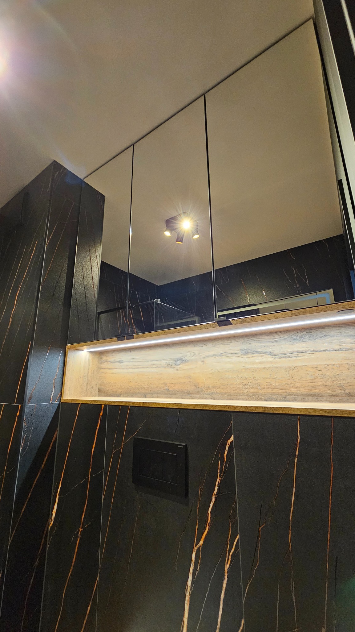 Elegancka łazienka z czarnymi płytkami imitującymi marmur, podświetlana półka z drewnianym dekorem i lustrzana szafka nad umywalką, widok z dołu.